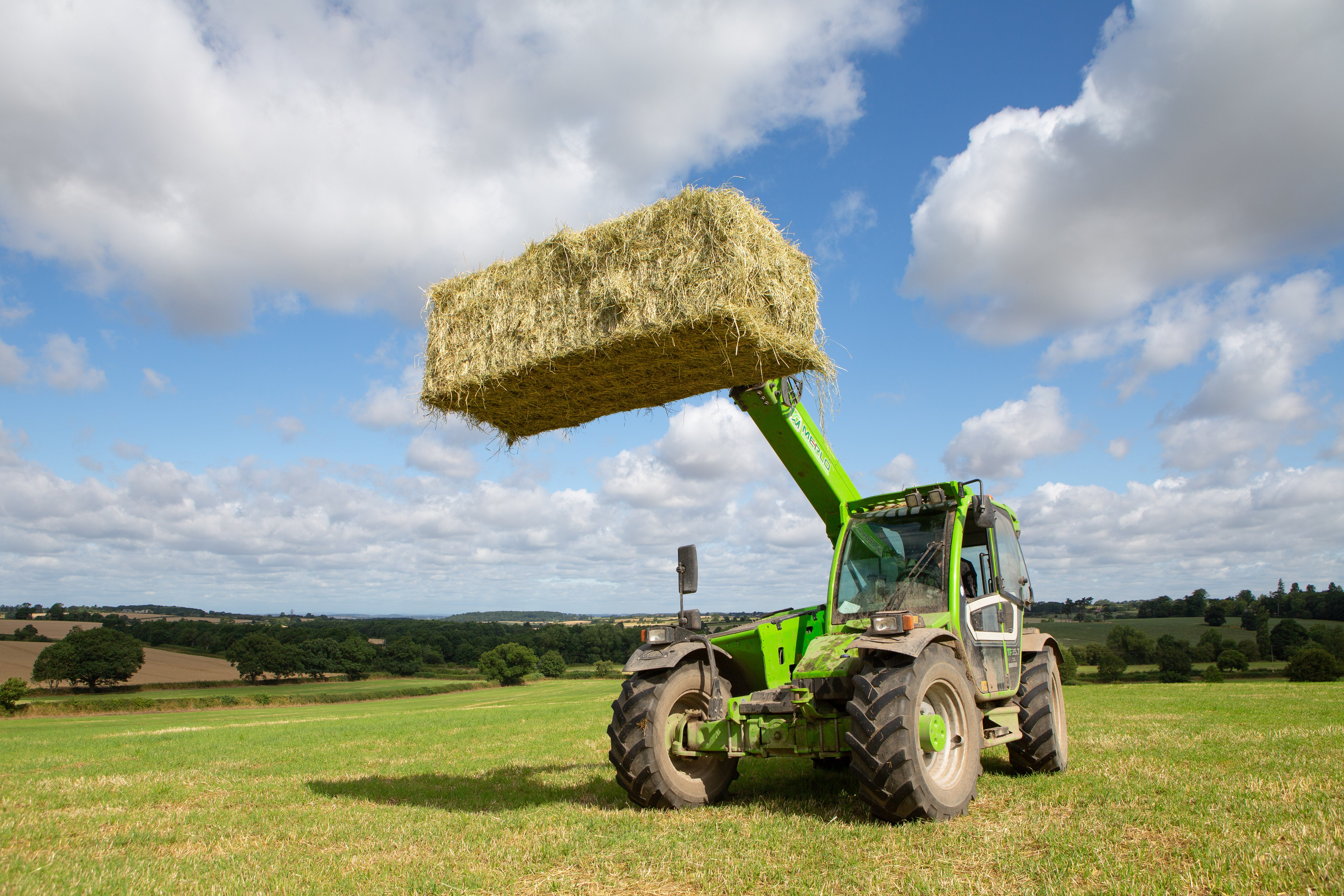 Green tractor lifting a bale of hay in an open field with a blue sky.