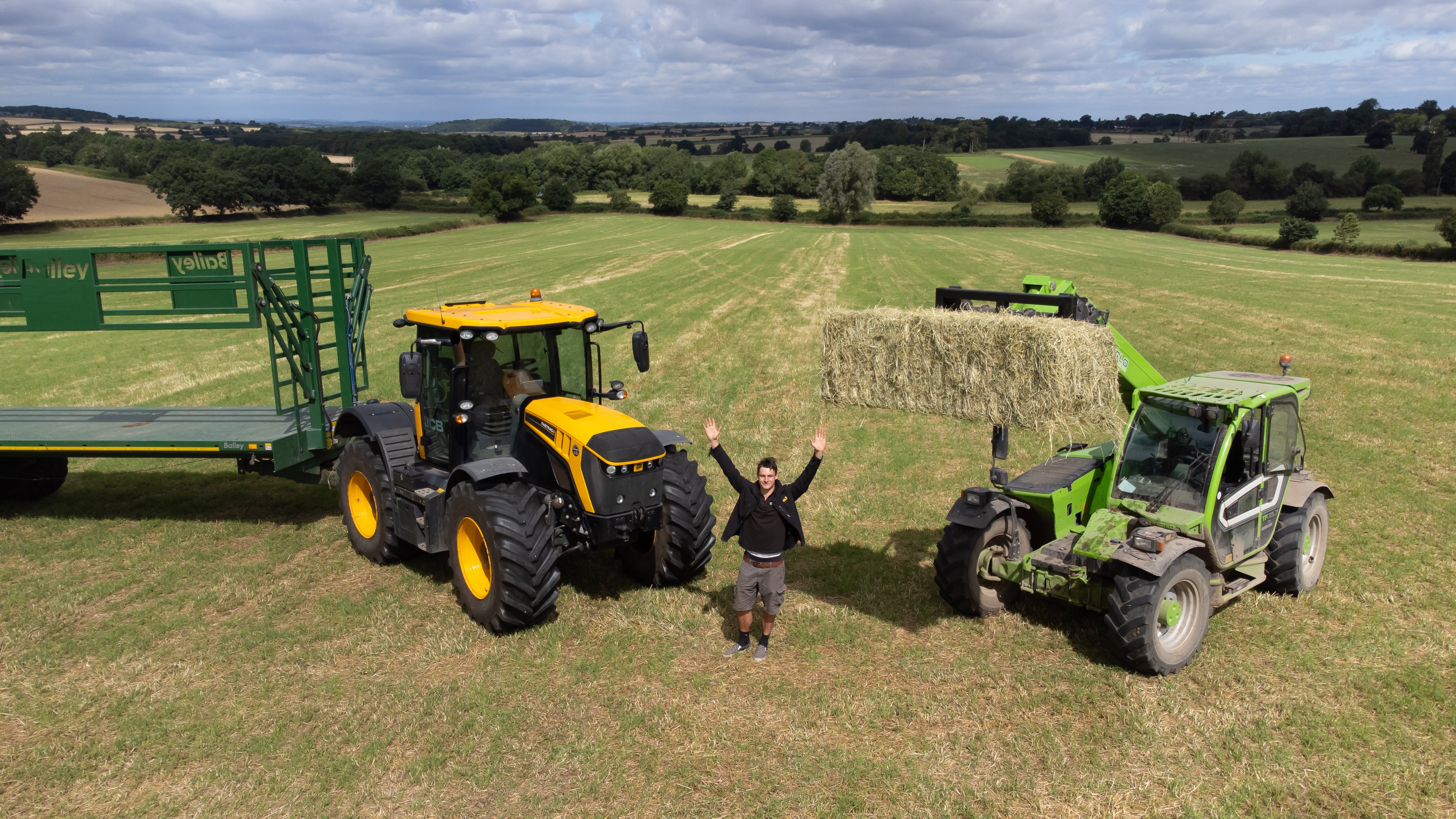 Person standing between a yellow tractor and a green telehandler on a farm field with hay bales.
